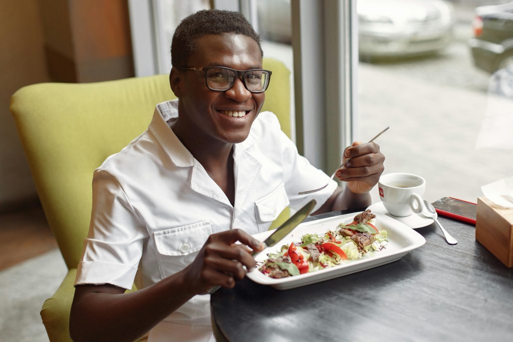 Smiling positive black man in stylish white shirt and eyeglasses eating delicious starter with mix salad leaves and meat slices while sitting at table in cafeteria near window